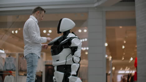 A-man-stands-with-a-robot-bot-and-asks-him-questions-and-asks-for-help-by-clicking-on-the-screen-on-the-robot-body.-Human-robot-interaction-in-the-modern-world