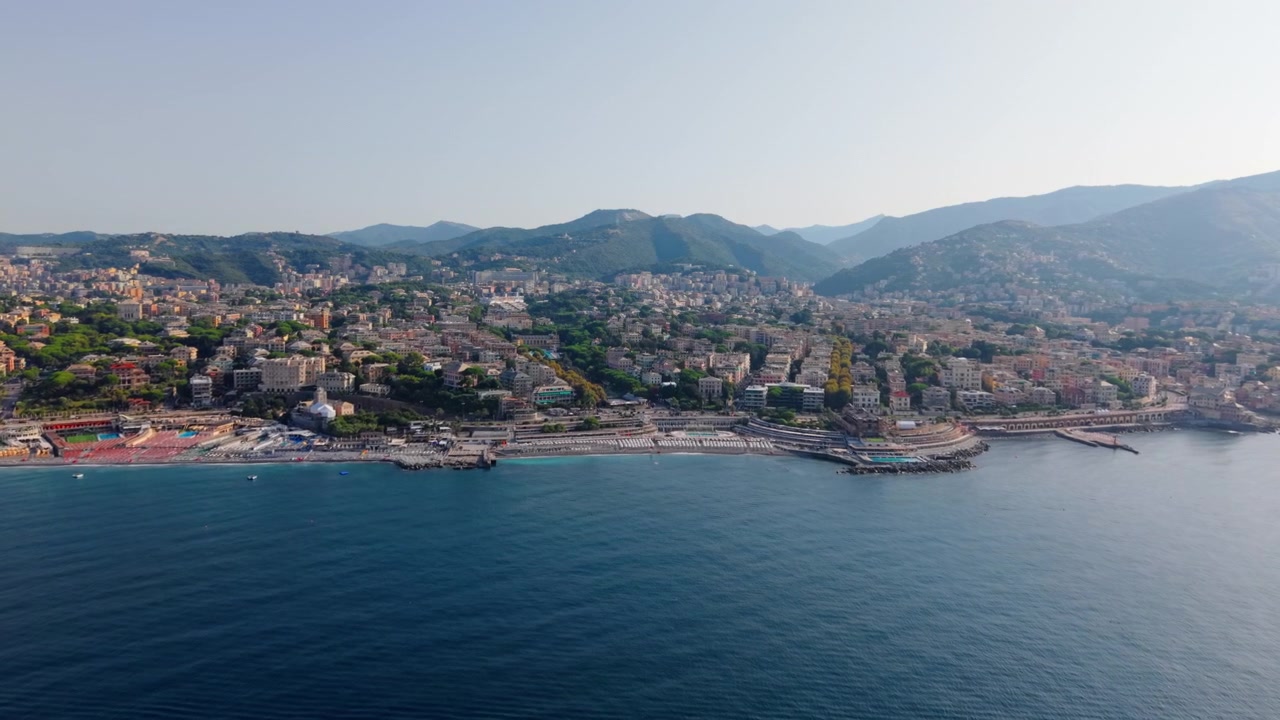 Vista aerea della costa di Genova con edifici cittadini e montagne in una giornata limpida.