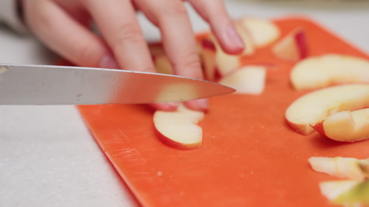 Close up hands slicing red apple into pieces with kitchen knife on orange plastic cutting board, crisp fruit prep, home cooking scene, safe technique, focus on detail