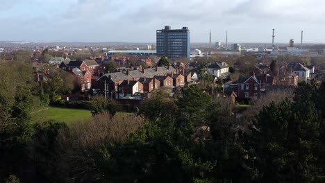 Aerial-view-over-park-trees-to-suburban-industrial-townscape-with-blue-skyscraper,-Merseyside,-England