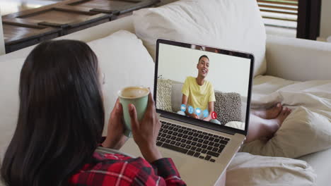african american woman holding coffee cup having a video call on laptop sitting on the couch at home