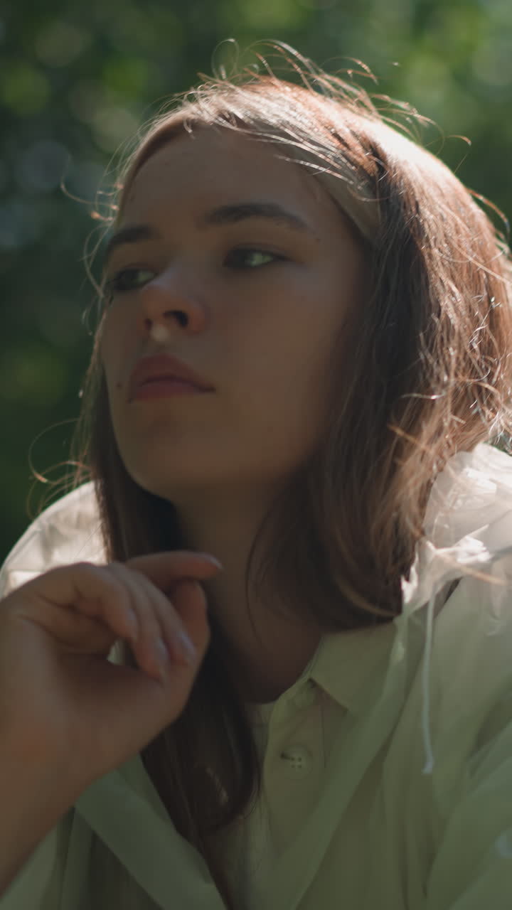 joven con impermeable translúcido baja la capucha y descansa la cabeza sobre la mano pensativamente, con la luz del sol reflejándose suavemente en su cara y la vegetación exuberante en el fondo