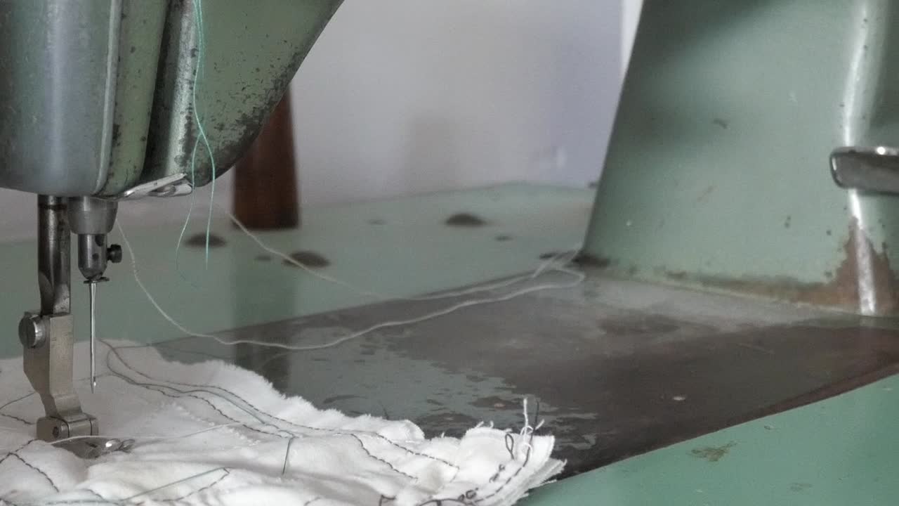 Pan right shot of a vintage Refrey sewing machine stitching white fabric with loose green thread in a workshop