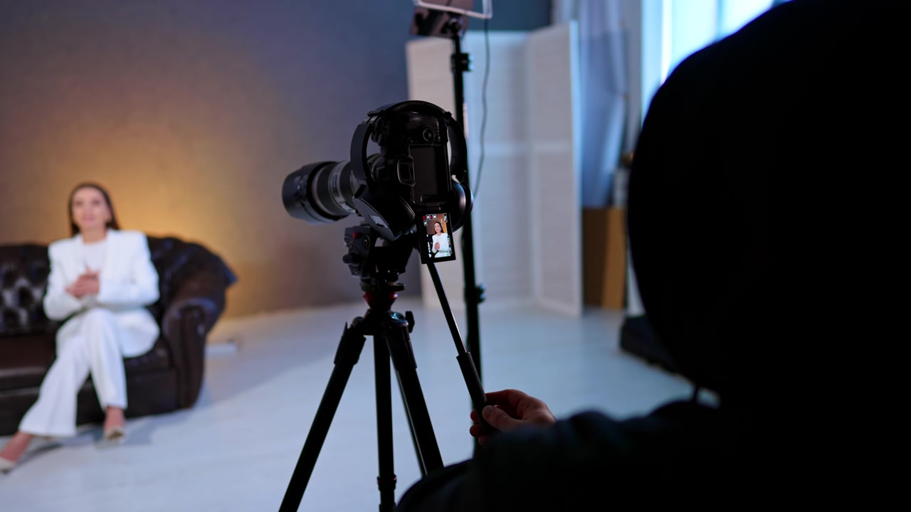 Kamera optager interviewperson i studieopsætning. Kamera på stativ filmer siddende kvinde, med sløret motiv, LED-lys og operatørs silhuet