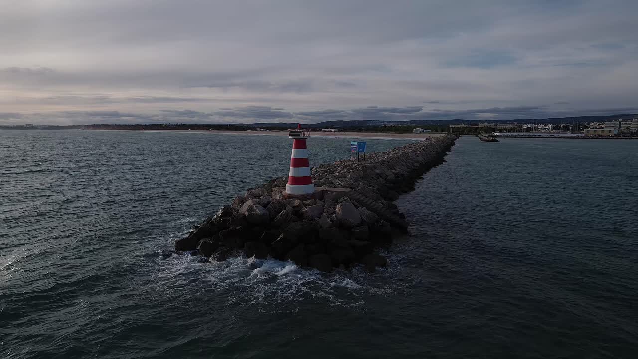 Drone orbit around the red‑and‑white lighthouse on the rocky breakwater at the entrance channel to Marina de Vilamoura,
