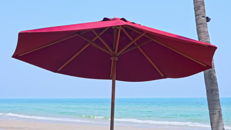 Toma-En-ángulo-Bajo-De-Una-Sombrilla-Roja-En-Un-Cielo-Azul-Claro-Con-Olas-Oceánicas-Que-Llegan-A-La-Orilla