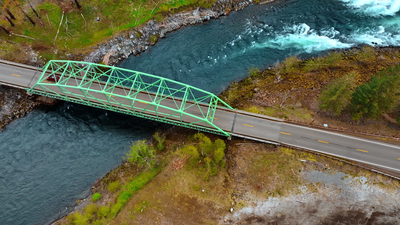 Red car rides by the road and crosses the bridge over quick river. Automobile travel in the mountainous area. Aerial view. Oregon wilderness.