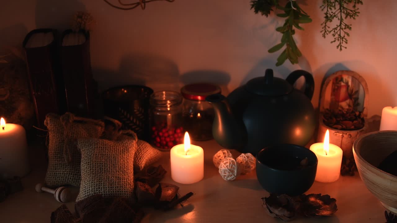 Scatto di dettaglio rilassante in sottofondo di un'erboristeria, con candele dalle fiamme tremolanti, una teiera, una tazza fumante, erbe appese, libri e della polvere che volteggia.