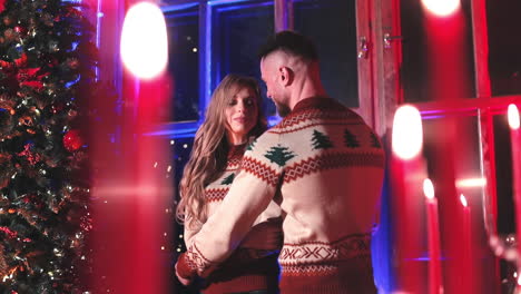 man hugging girlfriend near christmas tree. young couple in the same knitted christmas sweaters standing together in the house in the evening.