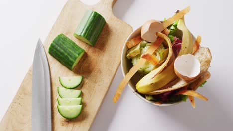 Overhead-video-of-knife-and-cucumber-on-chopping-board,-with-organic-waste-in-kitchen-composting-bin