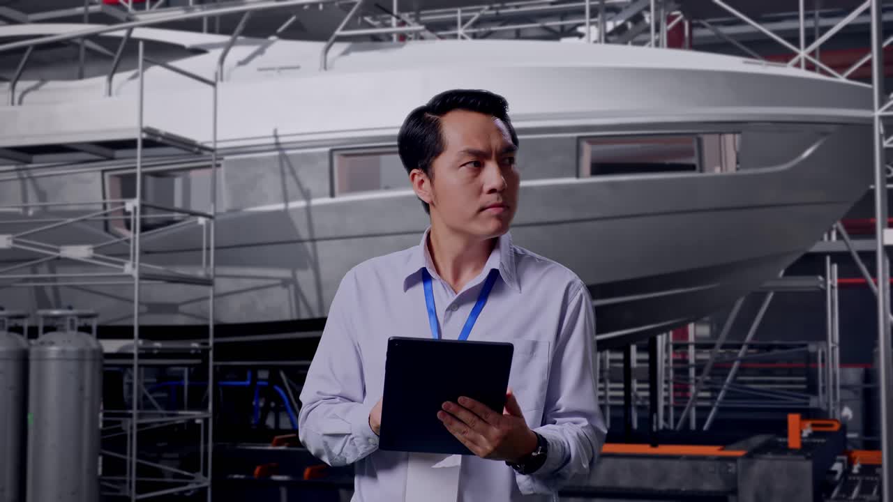 Asian Male Professional Worker Standing With His Tablet with Yacht Construction in Modern Shipyard, He Observes By Looking Around Before He Come To Concentrating With His Tablet