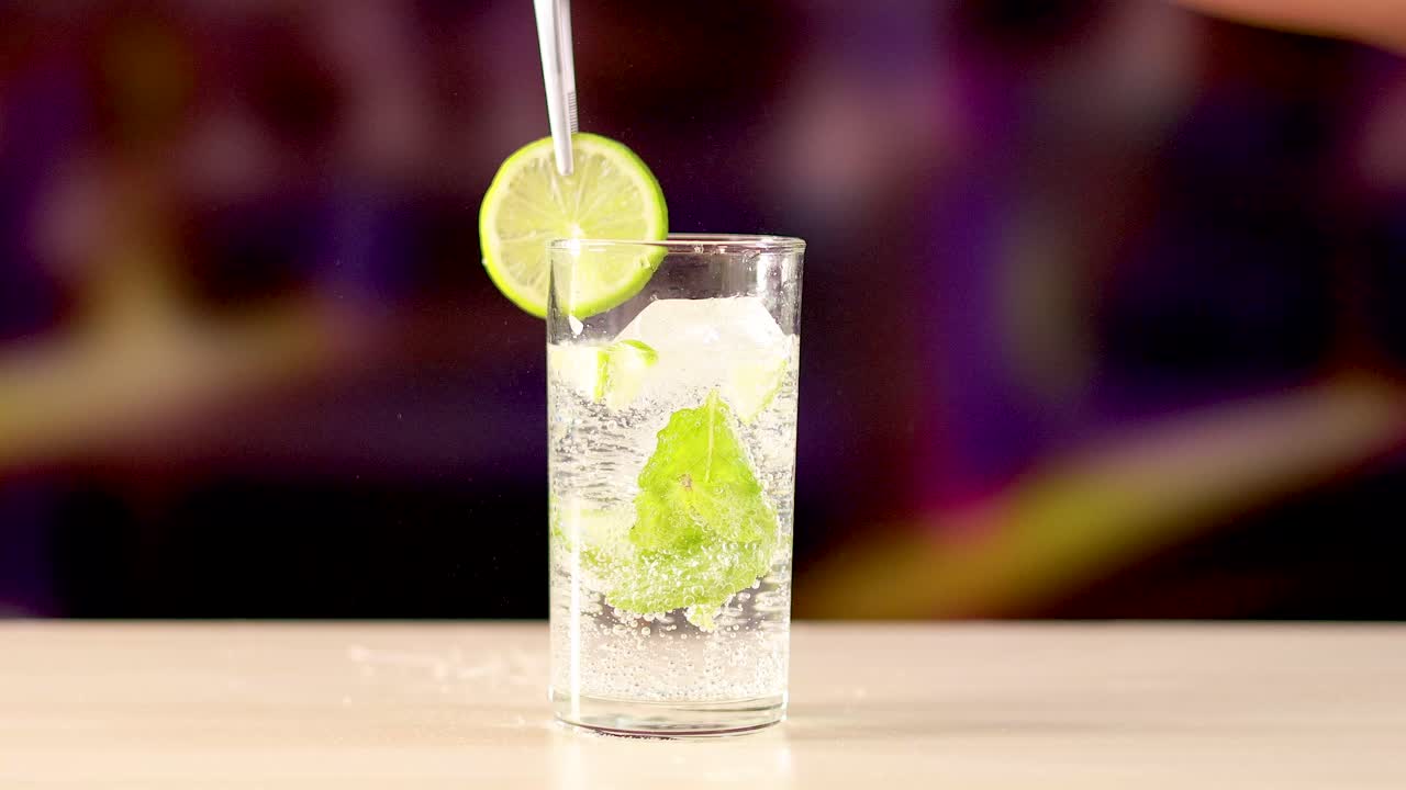 A lime slice is added to a mojito cocktail in a vibrant bar setting. Bright lighting enhances the refreshing drink