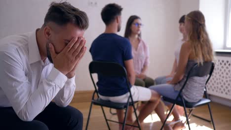 group therapy session, sad man covers his face with hands on background of people sitting on chairs in circle