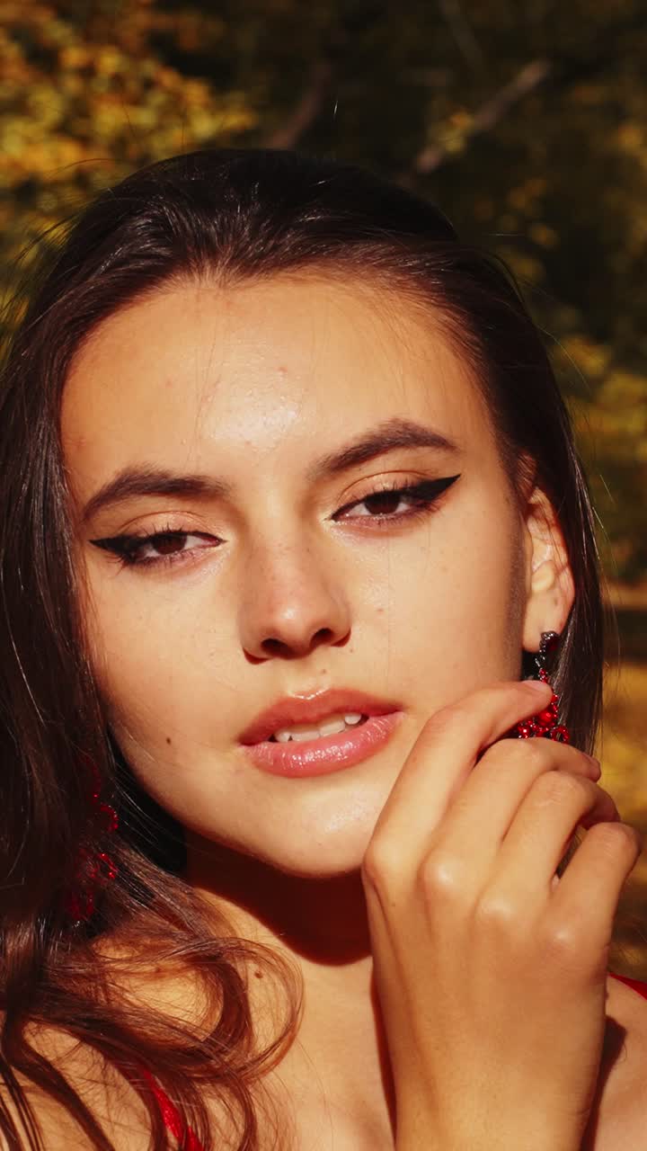 Ritratto affascinante di una giovane donna con trucco sorprendente ed eleganti orecchini rossi, sullo sfondo di un rigoglioso fogliame autunnale