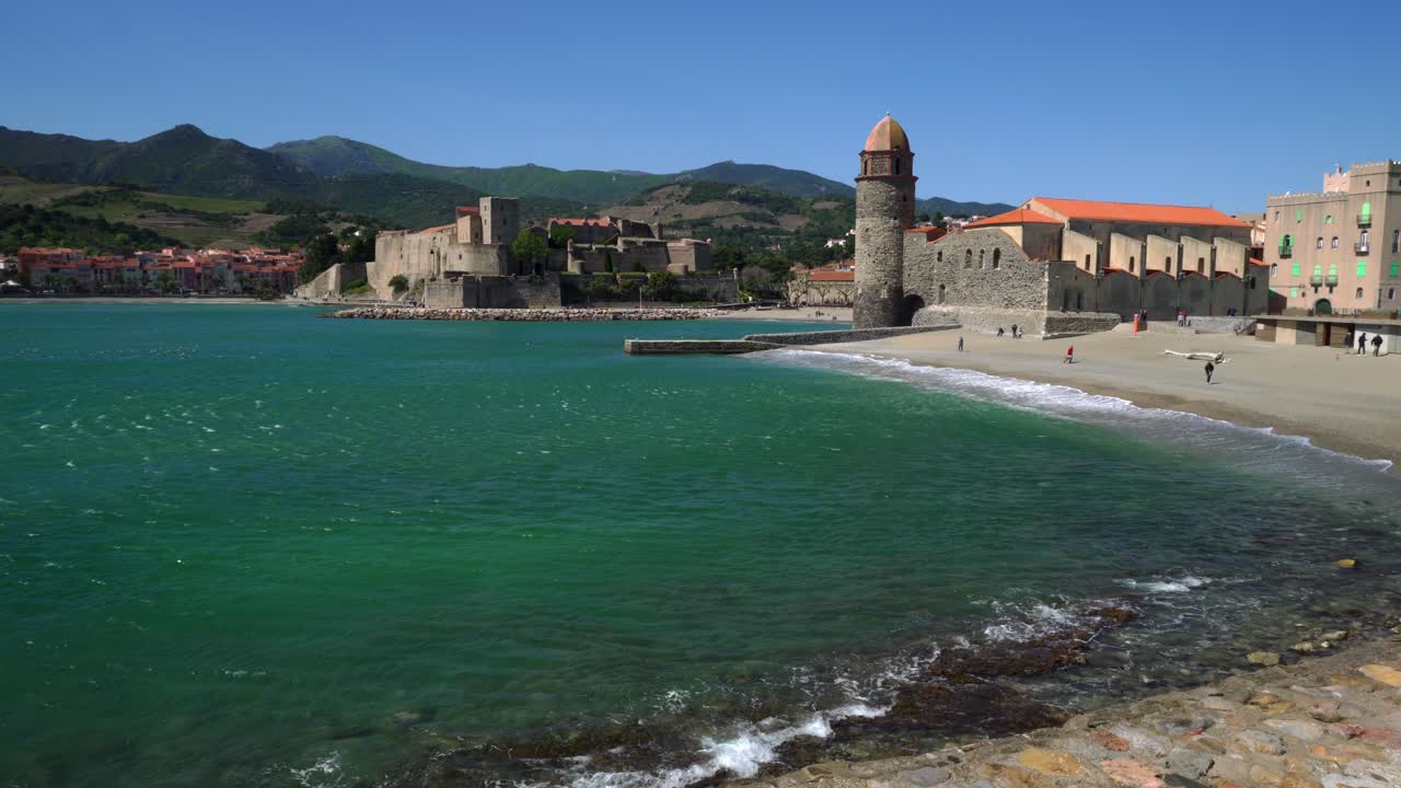 Onde che si infrangono sulla spiaggia durante forti venti a Collioure, con la città storica sullo sfondo.