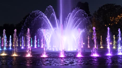 Fuente-Iluminada-Por-La-Noche-En-El-Parque-De-La-Ciudad,-Luces-Abstractas-Con-Colores-Cambiantes-De-Múltiples-Boquillas-En-Círculo