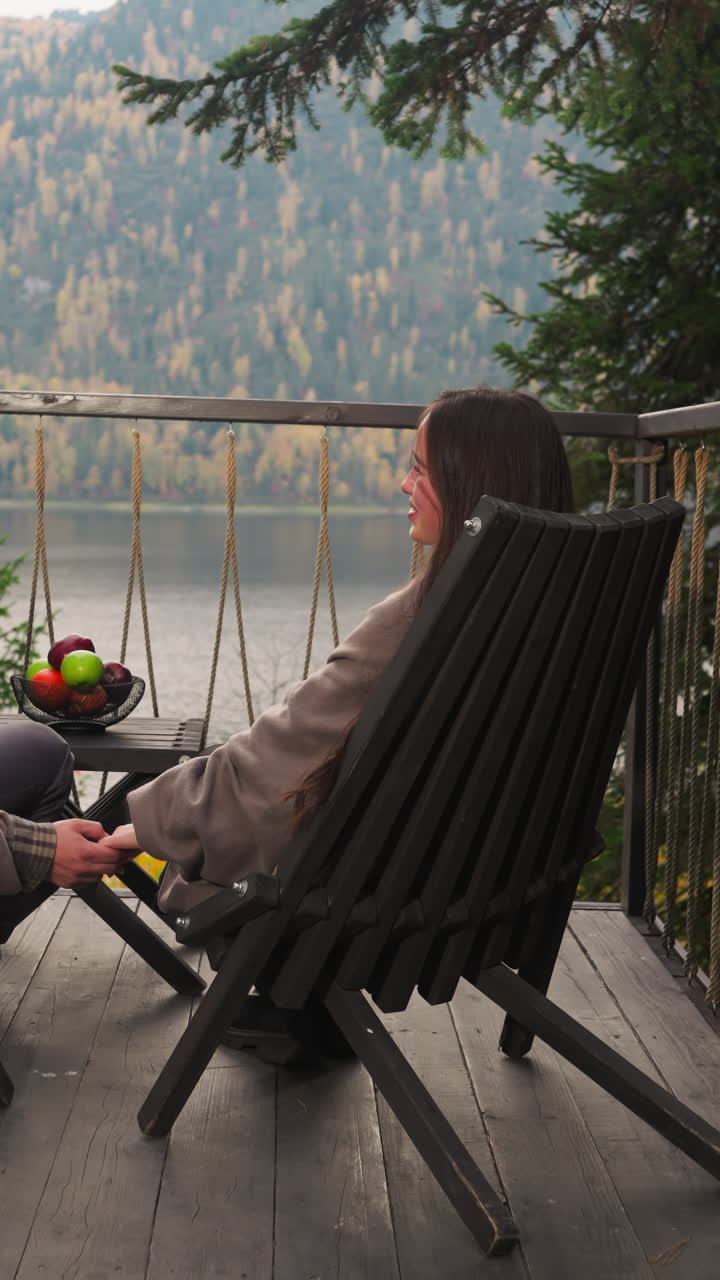 Husband pets wife hand sitting in armchairs. Woman talks to man on hotel terrace holding hands against river and mountains. Eco-friendly glamping resort