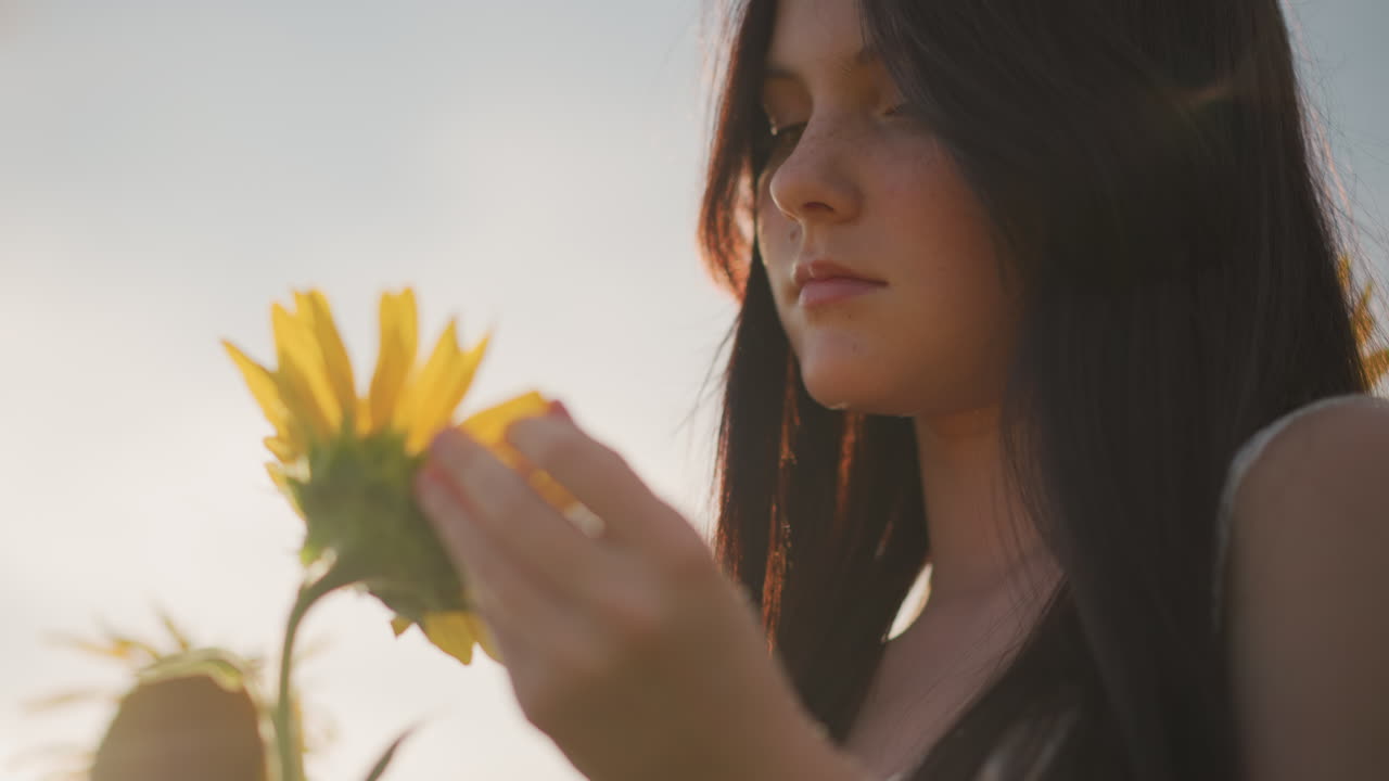 mujer caucásica joven en un campo de girasoles al atardecer, luz dorada suave iluminando el rostro desde atrás, perfil contemplativo, tocando los pétalos con delicadeza, atmósfera serena de pradera, ambiente cinematográfico en primer plano