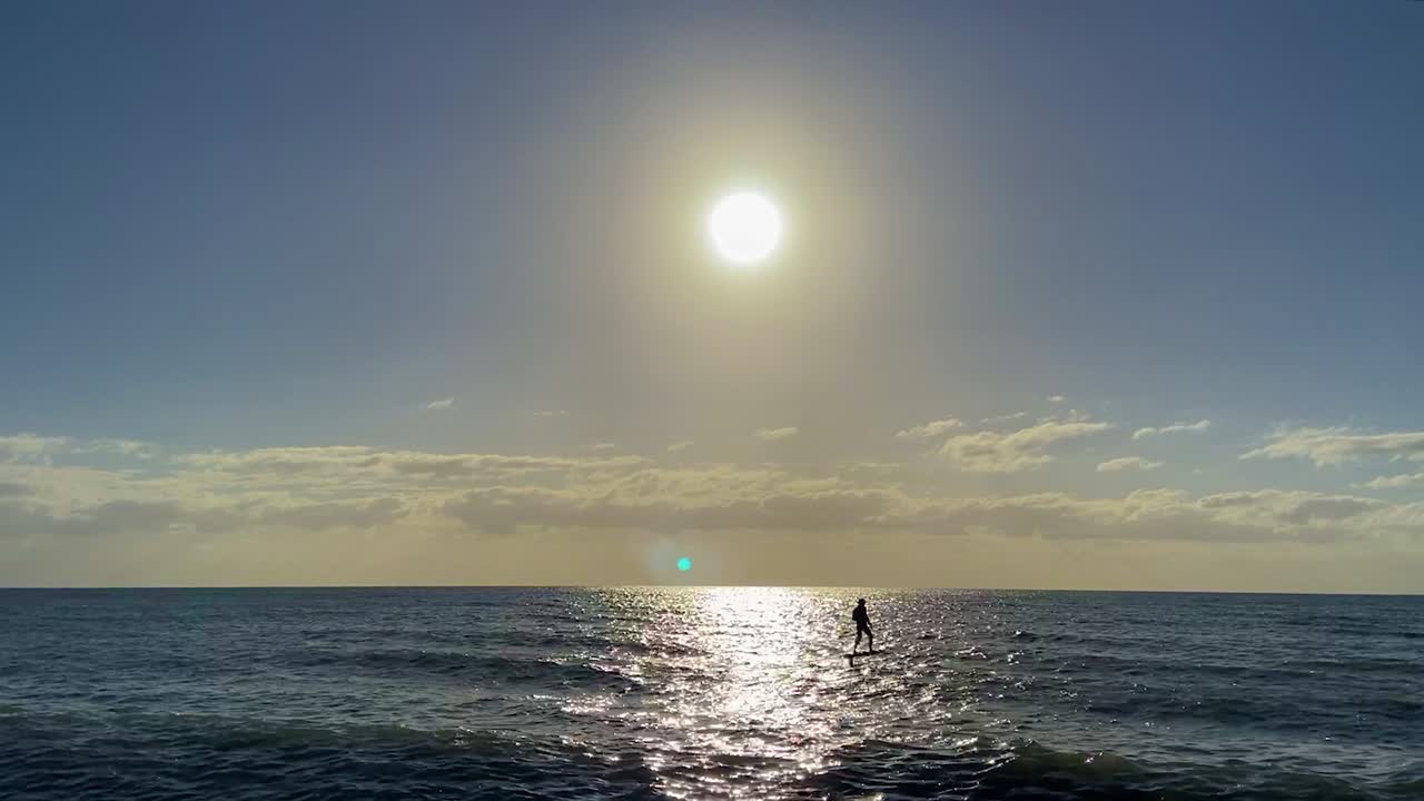 Stunning sunset over a tranquil tropical ocean, with a figure cruising past in silouette on a hydrofoil surf board, as the sun sparkles off the flat calm ocean