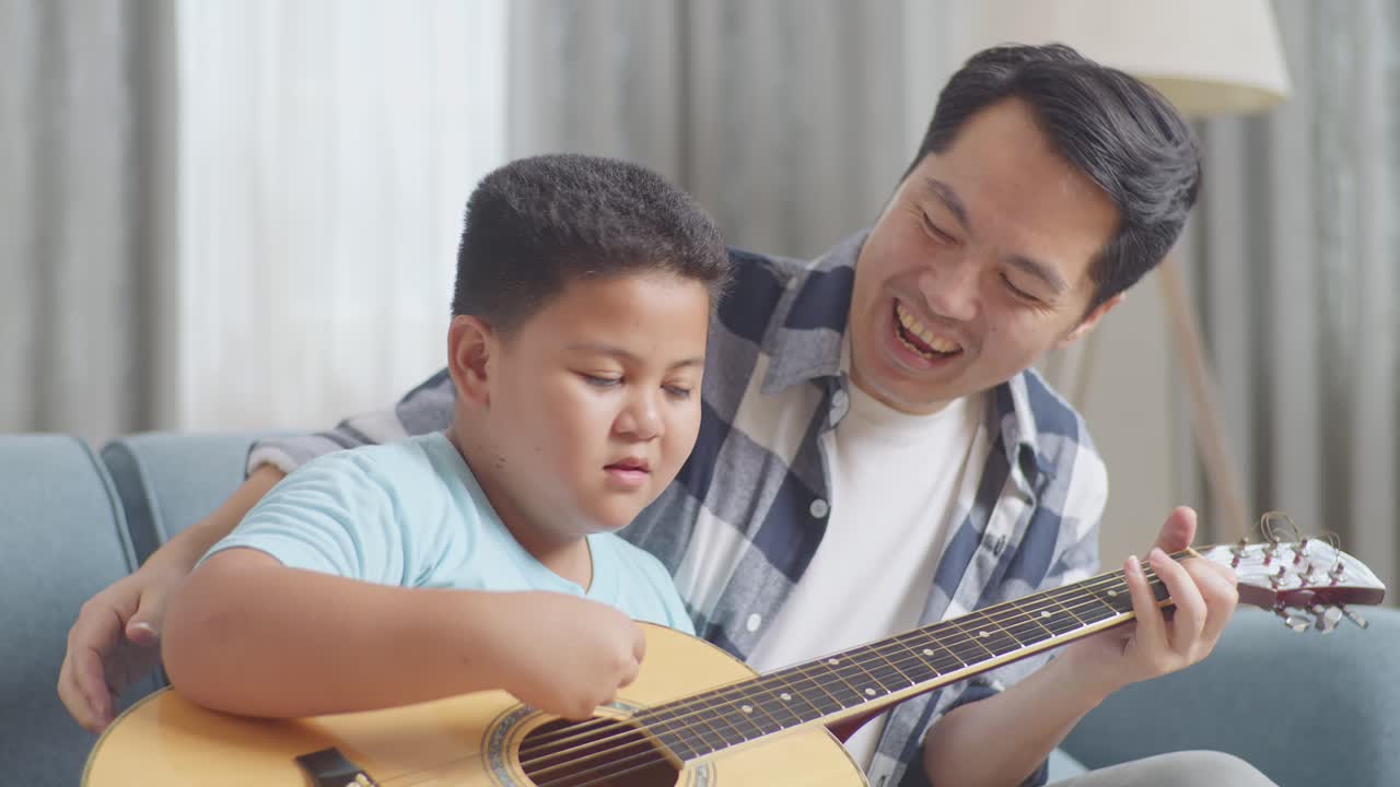 幸せなアジアの父と息子が自宅のソファで一緒にギターを弾くことができることを祝う