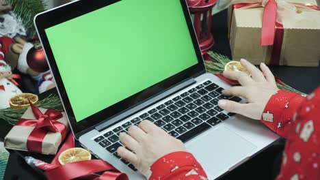 woman is using laptop with green screen in front of decorated christmas tree. fingers are scrolling news, watching photos in social media, typing messages. chroma key. online christmas shopping