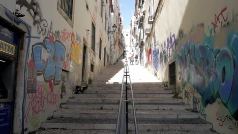 very steep street in lisbon with steps only, calcada da bica grande. long handrail in the middle. graffiti painting on walls of buildings. many windows some drying clothes hanging. an atm machine.