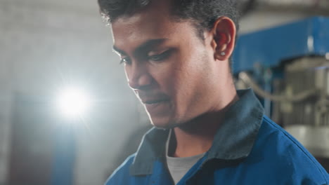 hombre en uniforme azul sonriendo mientras se centra en algo fuera de la cámara, iluminado por una luz brillante en un entorno de taller con equipos mecánicos visibles