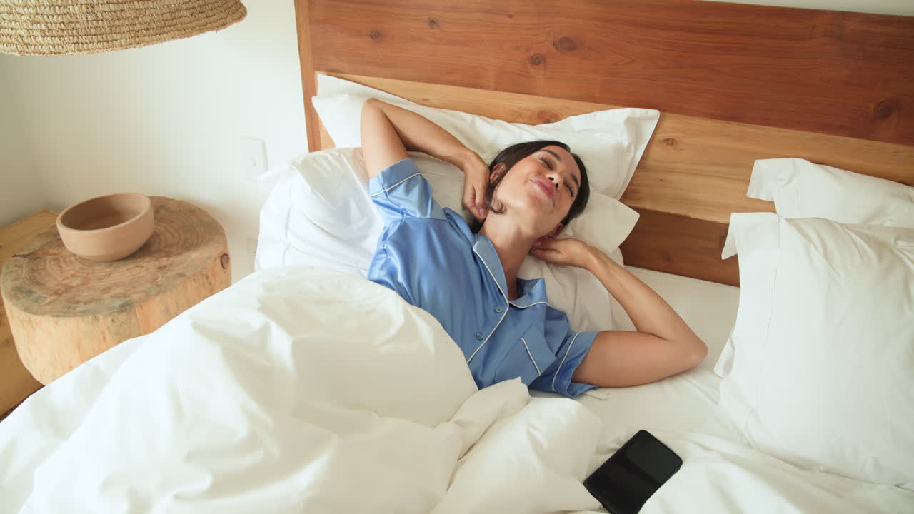 Woman in blue pajamas waking up in bed, smiling and stretching happily, at home