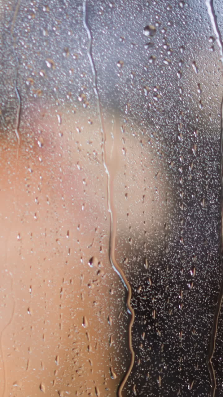 Steamy from condensation transparent glass shower booth wall with water drops flowing down against woman bare back in bathroom slow motion closeup