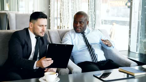 two multi-ethnic businessmen looking at clipboard and discussing startup projects in glassy cafe during lunch time