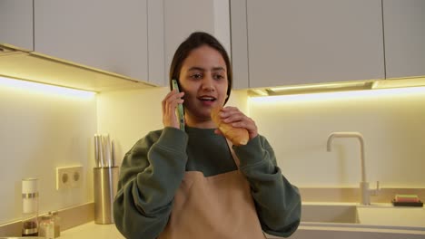 una chica morena feliz y segura en una chaqueta verde y un delantal beige come un croissant mientras se comunica con sus seres queridos usando un teléfono verde y se regocija en un apartamento moderno en la cocina