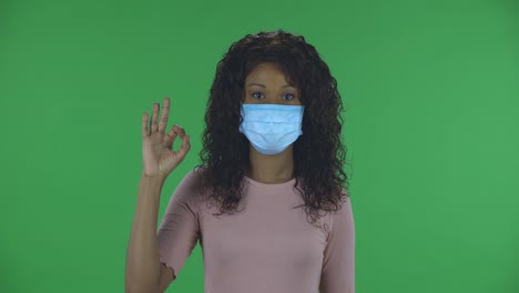 portrait of beautiful african american young woman in medical mask is looking straight sighs relieved and making sign ok. burning brunette with wavy hair in jeans and a beige blouse on a green screen in the studio. health protection corona virus concept