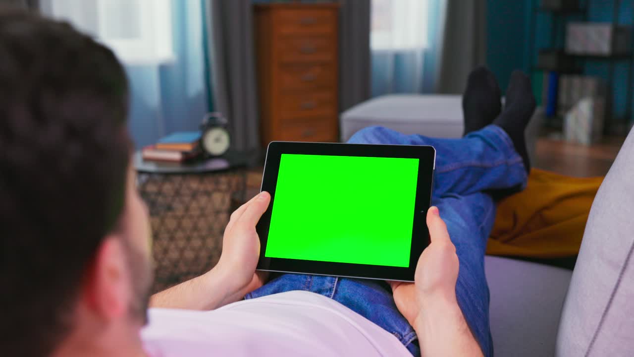 Young Man at Home Laying on a Couch Using with Green Mock-up Screen Tablet Computer. Man Using Touchscreen Device, Browsing Internet, Watching Content, Videos