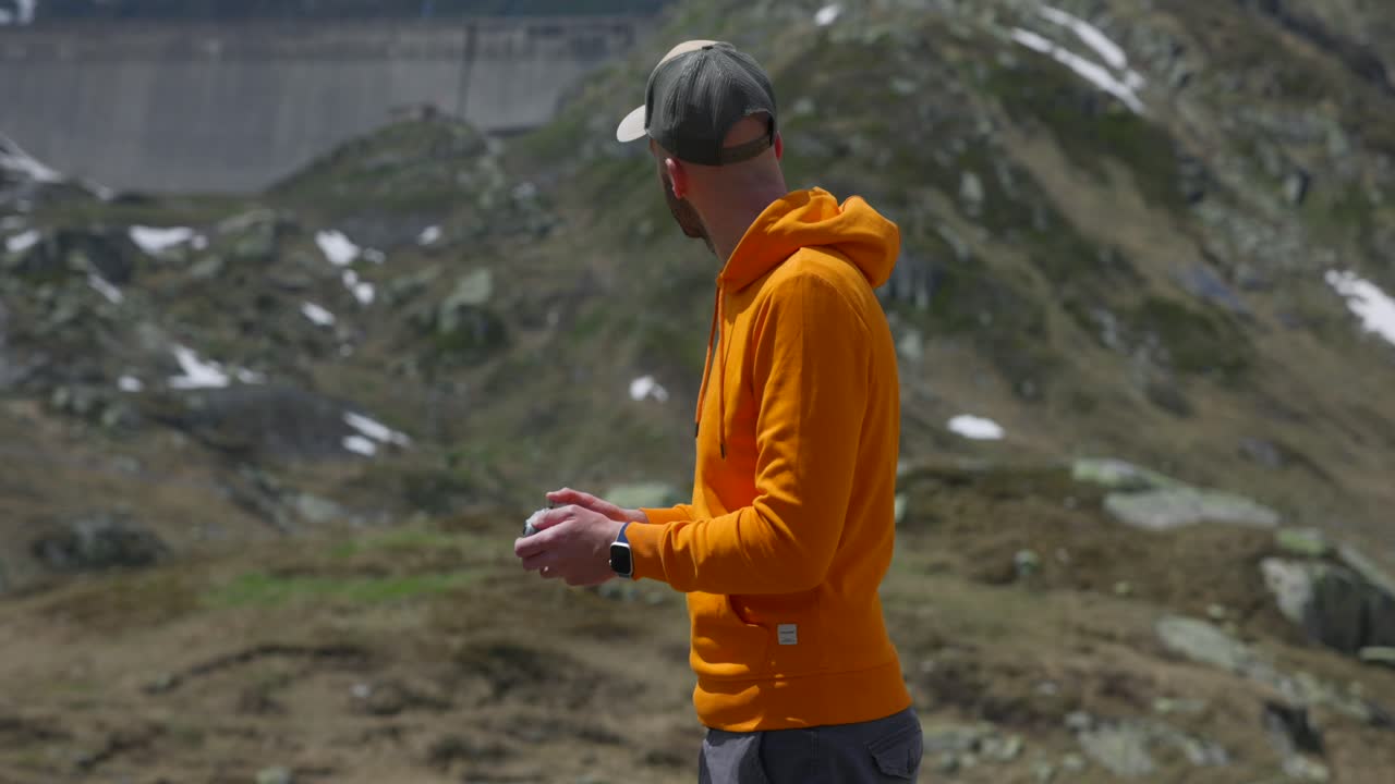 Adult Male Wearing Orange Hoody And Baseball Cap Looking At Drone Controller Unit As He Pilots Craft