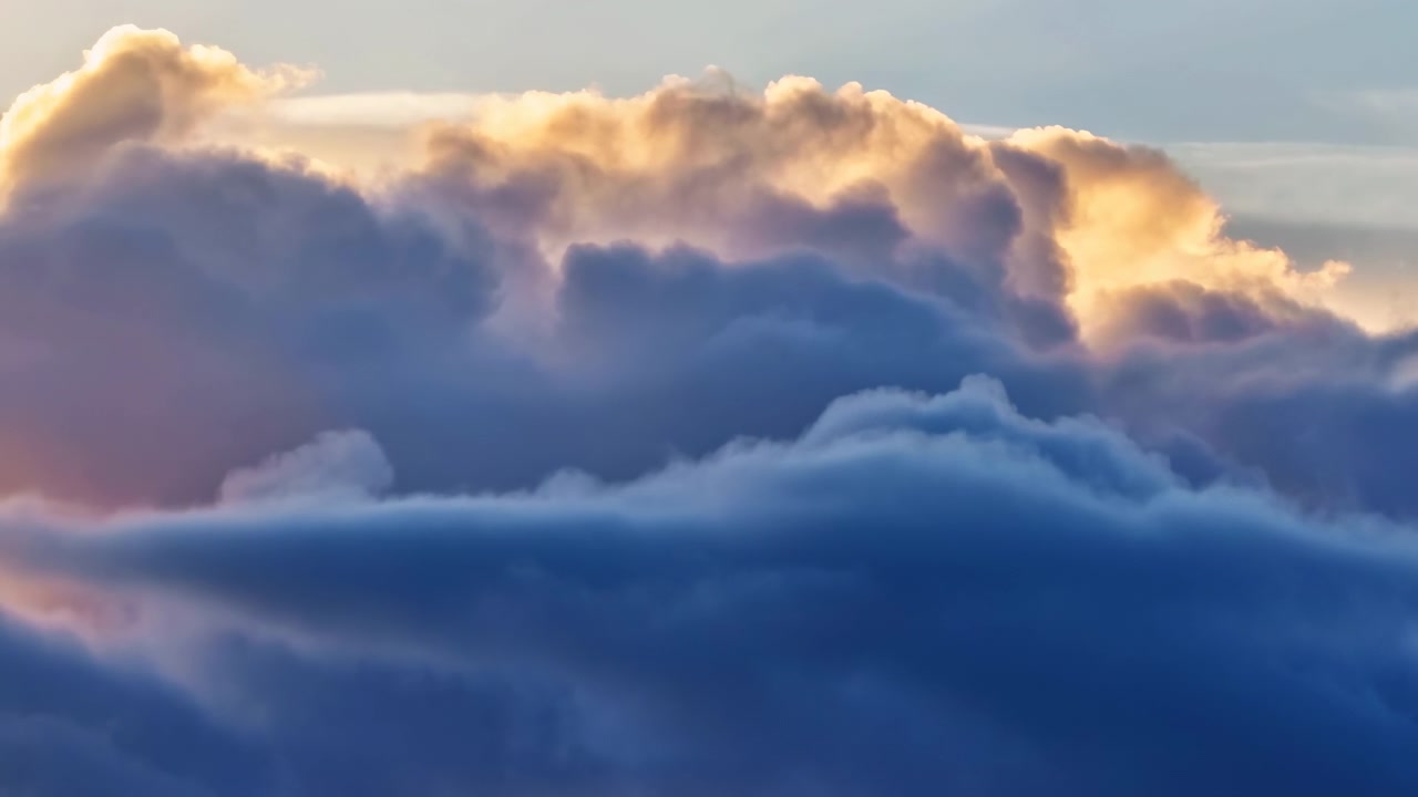 Imponenti strati di nuvole si rimodellano lentamente nell'inquadratura mentre una luce calda e bassa ne illumina i bordi superiori, contrastando i riflessi luminosi color crema con le profonde masse d'ombra blu-grigio.