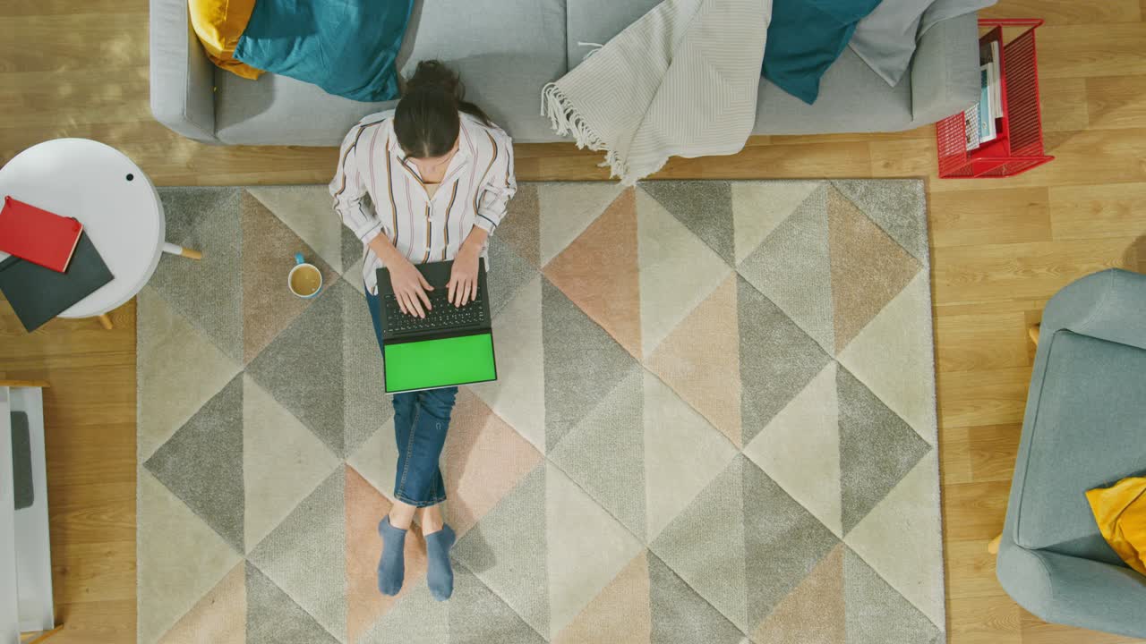 una joven está sentada en un piso, bebiendo café y usando una computadora portátil con pantalla verde. acogedora sala de estar con interior moderno con alfombra, sofá, silla, mesa, estantería, planta y piso de madera. vista superior con zoom out.