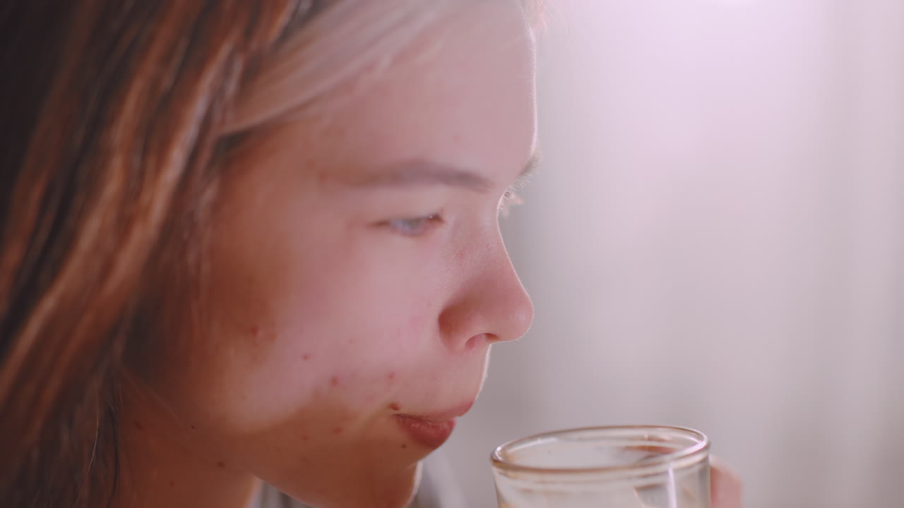 Close up of young lady holding sandwich in hand while sipping tea from glass cup, soft indoor light reflecting on drink, capturing calm relaxed lifestyle moment of nourishment