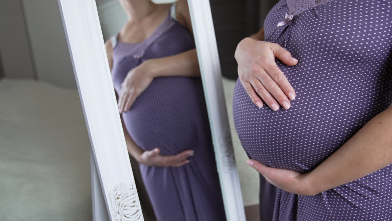 Expecting white woman strokes tummy in front of the mirror. Pregnant lady preparing for a childbirth. Expectant mom in a pajama in her comfortable bedroom.