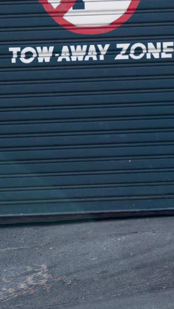 Video verticale: Uomo che fa jogging tiene una tazza trasparente, persone passano davanti a una saracinesca con la scritta "ZONA RIMOZIONE FORZATA".