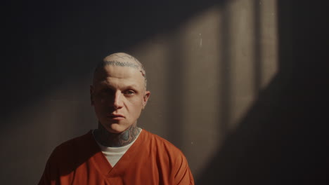 portrait of bald tattooed inmate in orange uniform sitting in prison cell