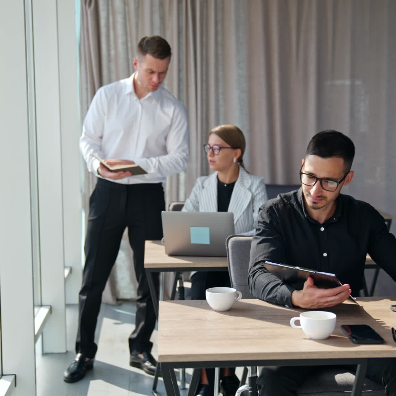 Office work in the light room. Male manager stands near female employee showing her paper notebook. Man at the foreground looking through the documents