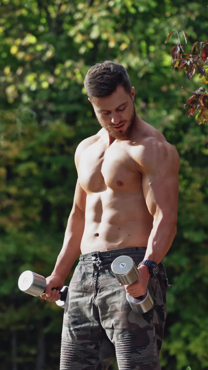 Bearded shirtless man doing workout in nature. Muscular bodybuilder exercising with dumbbells outdoors. Handsome athlete lifting dumbbells. Vertical video