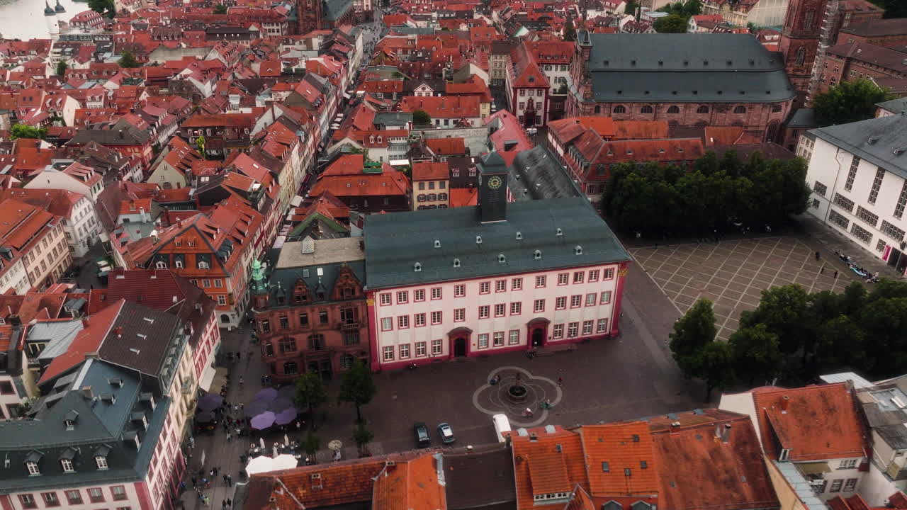 The drone flies backward above Heidelberg’s old town, showing dense red rooftops, narrow streets, a large church and surrounding buildings as it arcs slightly across the cityscape
