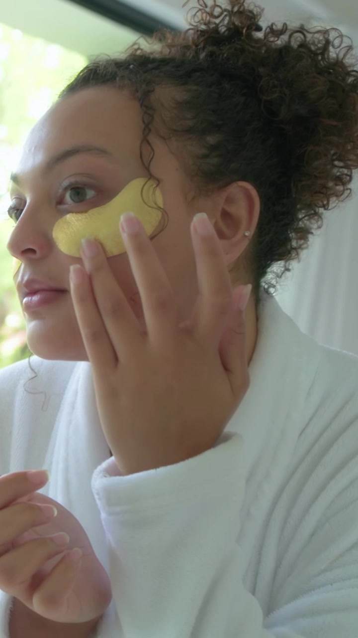 Vertical video: Holding gold under-eye patch, woman applying patches in robe at mirror for skincare