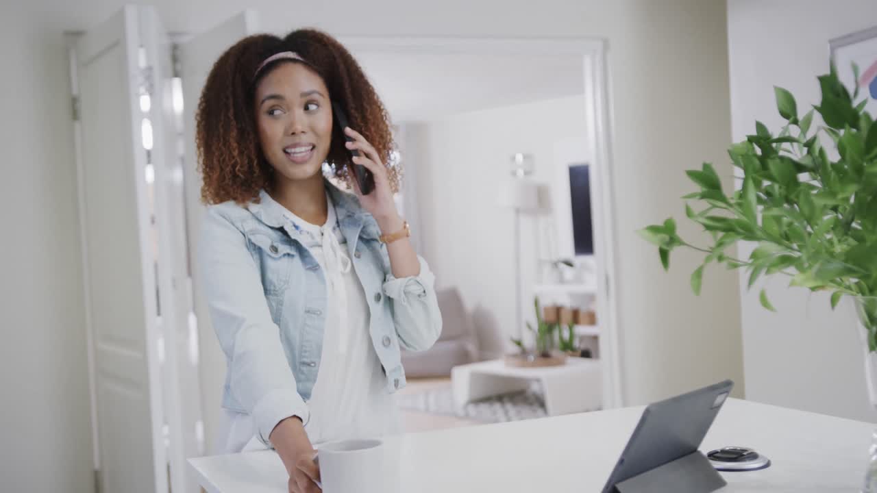 Happy biracial woman talking on smartphone having coffee in kitchen with tablet, slow motion