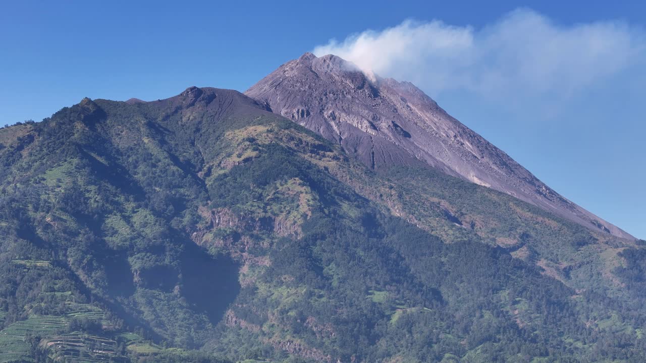 Aerial view of scenic landscape of smoking volcanic peak with forest-covered slopes and vibrant greenery in bright daylight. Volcanic mountain of Merapi, Indonesia
