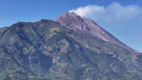 Aerial view of scenic landscape of smoking volcanic peak with forest-covered slopes and vibrant greenery in bright daylight. Volcanic mountain of Merapi, Indonesia
