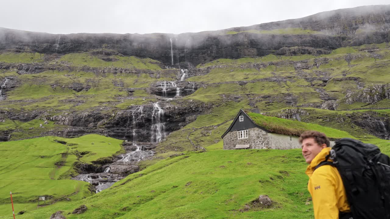 A man hiking toward a remote grass-roofed house in the misty green hills of Saksun