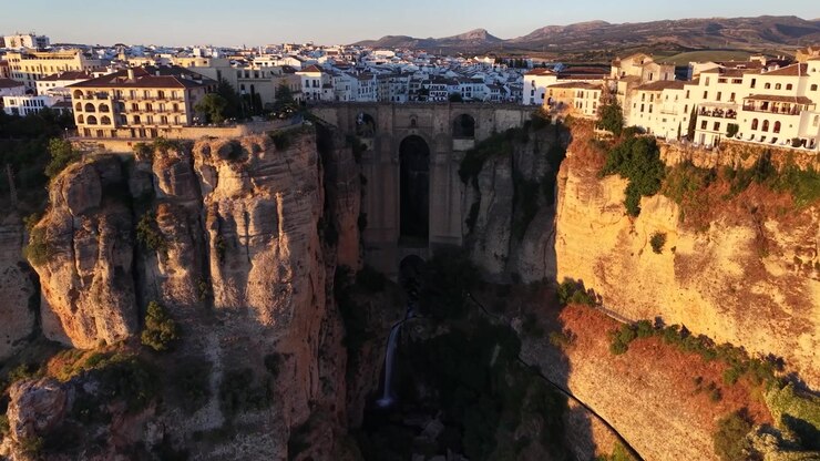 Wide drone aerial view of Puente Nuevo Bridge spanning El Tajo Gorge in Ronda, Spain during golden hour with dramatic cliffs and historic whitewashed buildings