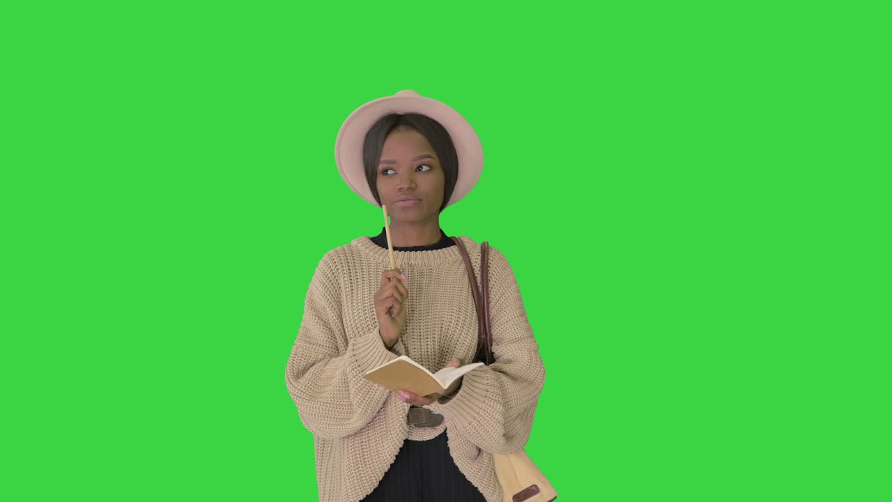 bonita mujer afroamericana en suéter tejido y sombrero escribiendo en su bloc de notas en una pantalla verde, clave de croma